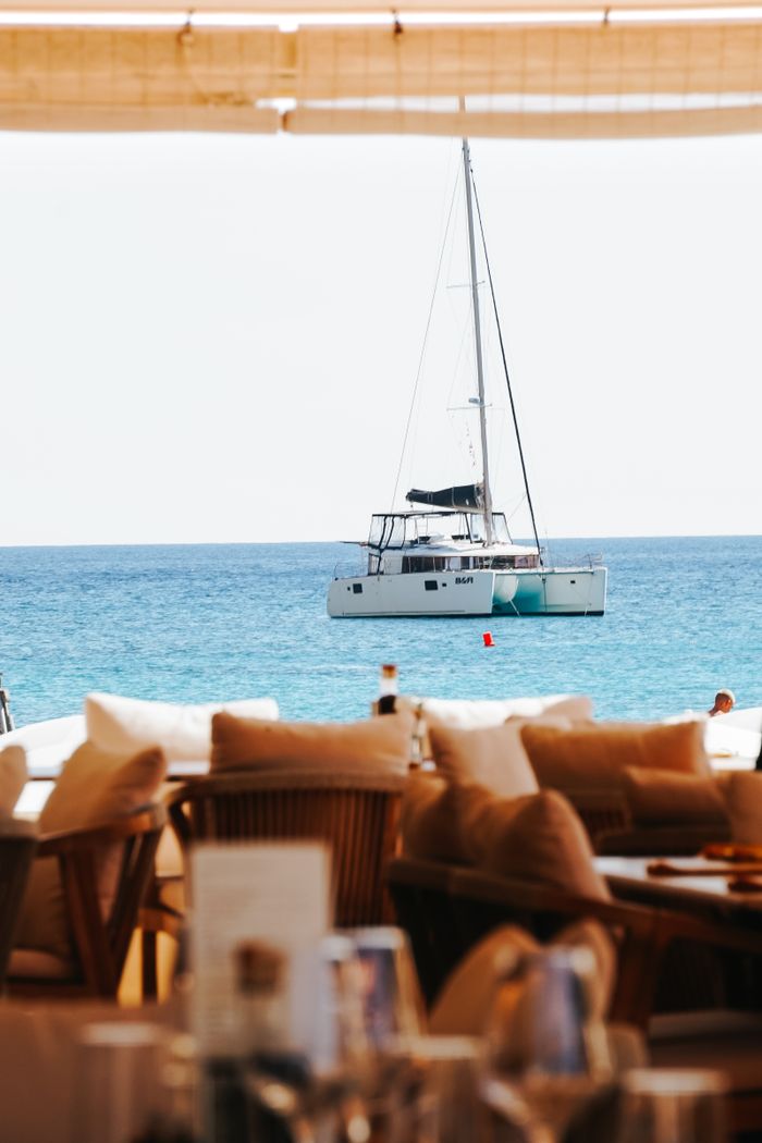 services-01 Sailboat in blue sea seen from cozy seaside restaurant terrace.