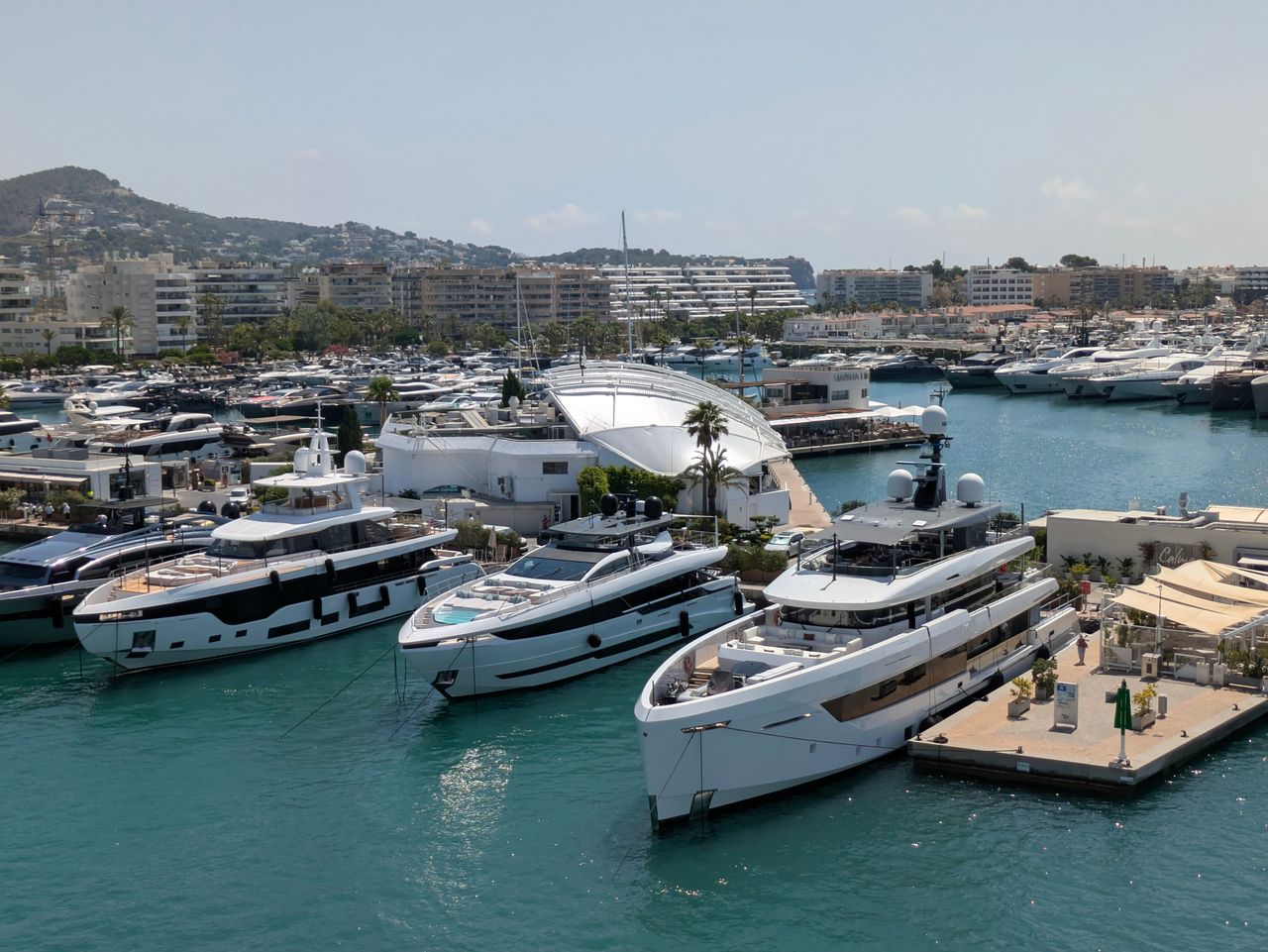 services-02 Aerial view of luxury yachts docked in a sunny Mediterranean marina, surrounded by hills and cityscape.